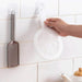 A white kitchen utensil is being hung on a transparent adhesive hook on a white tiled wall. Next to it, a metal grater is also hanging on a similar hook.