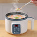 A white multi-functional cooker with digital controls, steaming hot soup. A hand is holding a spoon with soup above the pot.