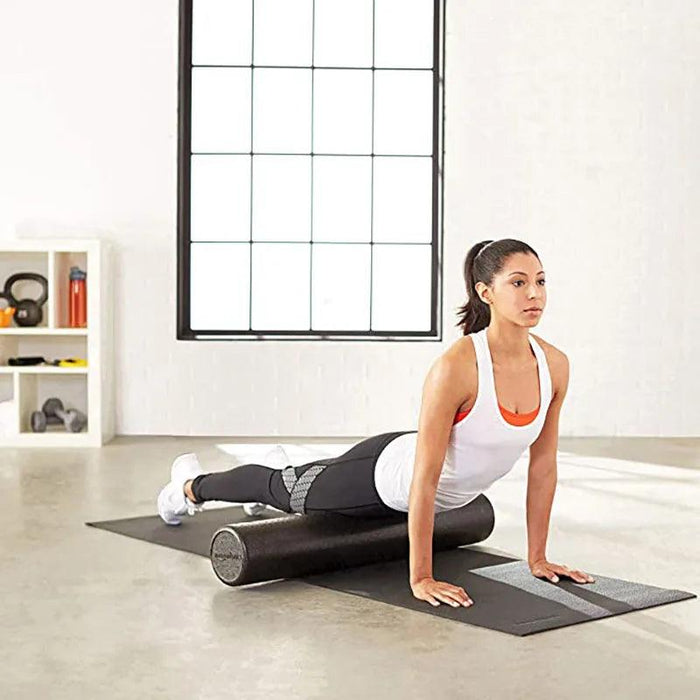 A woman using a black foam roller for exercise on a yoga mat in a bright room.