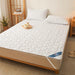 A white quilted mattress cover pad on a bed, with matching pillows. The room has a cozy setup with a small table, a plant, and slippers beside the bed.