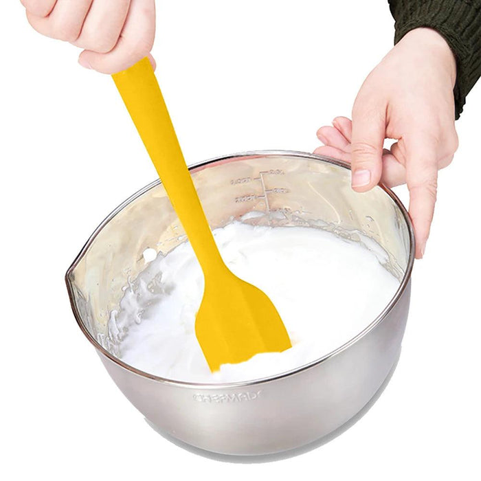 A yellow silicone spatula is being used to mix a white batter in a stainless steel bowl. The spatula is flexible and has a wide, flat head, making it ideal for mixing and scraping.