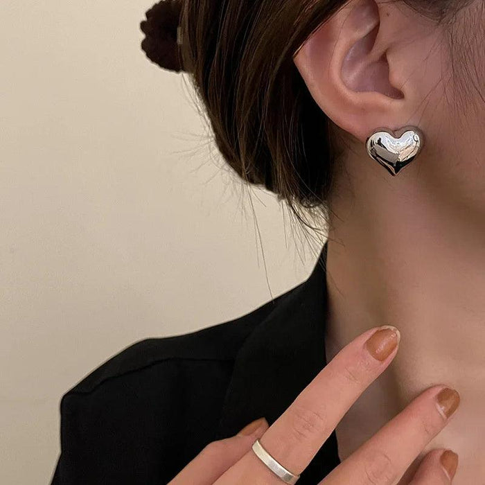 a woman wearing silver heart-shaped stud earrings. The earrings have a smooth, polished surface, giving them a sleek and elegant appearance. She is wearing a black top, which contrasts nicely with the shiny earrings.