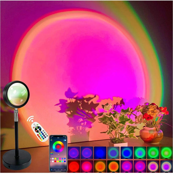 This image highlights the lamp's ability to create a vibrant and colorful atmosphere in a room. The lamp projects a large pink and purple gradient circle on the wall, with plants and flowers in the foreground. The photo also shows a remote control and smartphone app, indicating remote control options.