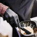 A close-up image of a hairdresser's hands wearing black gloves while holding a comb and mixing hair dye. This image highlights the use of gloves for protection in hair and beauty services.