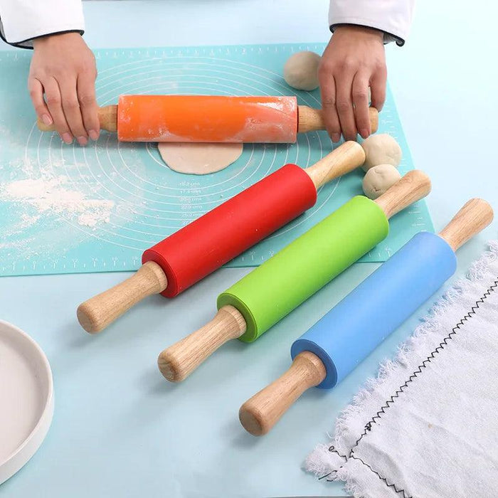 a person using an orange rolling pin to roll out dough on a blue silicone mat. Three other rolling pins in red, green, and blue are laid out on the same mat alongside small dough balls. The scene suggests a baking setup with a plate and a piece of cloth nearby.