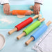 a person using an orange rolling pin to roll out dough on a blue silicone mat. Three other rolling pins in red, green, and blue are laid out on the same mat alongside small dough balls. The scene suggests a baking setup with a plate and a piece of cloth nearby.