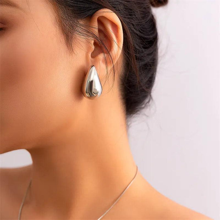 a woman wearing large, teardrop-shaped silver earrings. The earrings are smooth and reflective, creating a bold and sophisticated look. The woman's hair is styled in a simple updo, allowing the earrings to stand out.