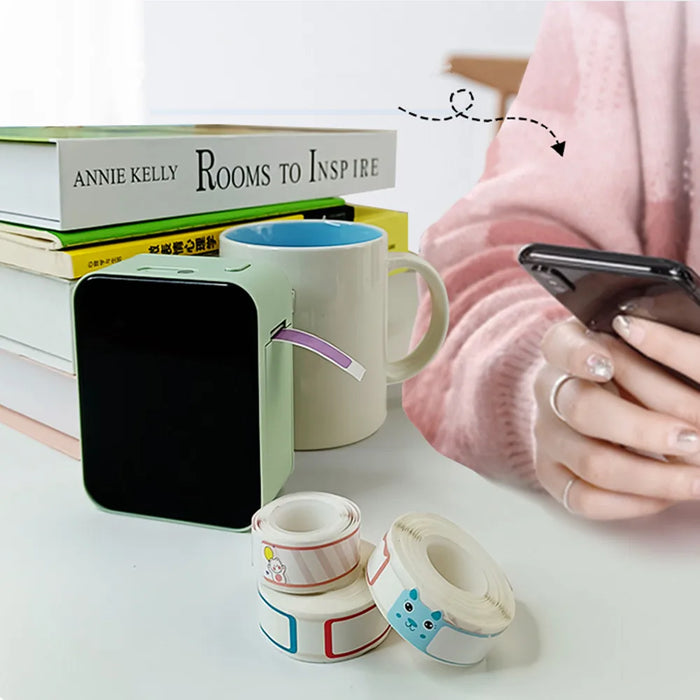 Green mini Bluetooth thermal label printer on a desk with a coffee mug, books, and rolls of decorative label tapes, next to a person using a smartphone.