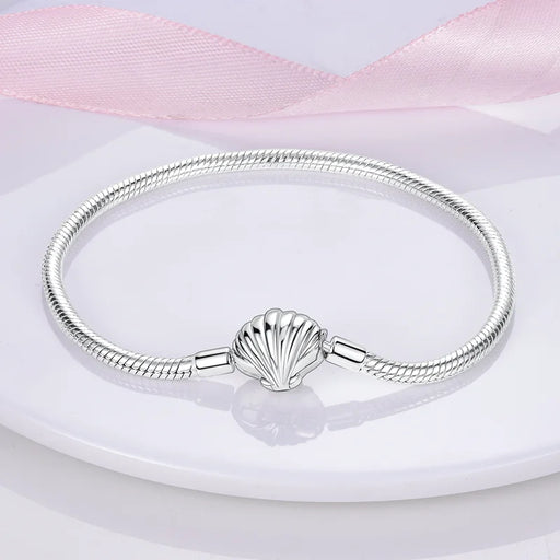 A silver bracelet with a seashell-shaped charm on a white background with a pink ribbon.