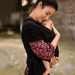 A mother tenderly holds her baby, who is comfortably nestled in a black baby sling and her shirt adorned with a colorful floral pattern. The mother's gentle smile and the secure embrace convey a moment of loving connection and protection, set against a soft-focus natural background that highlights the intimacy of the scene.