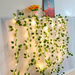 A festive indoor decor featuring a wall draped with a white cloth, decorated with cascading green ivy and twinkling lights. Above this setup, a shelf holds a colorful painting and a small decorative item, adding a touch of personal style and vibrancy to the space.